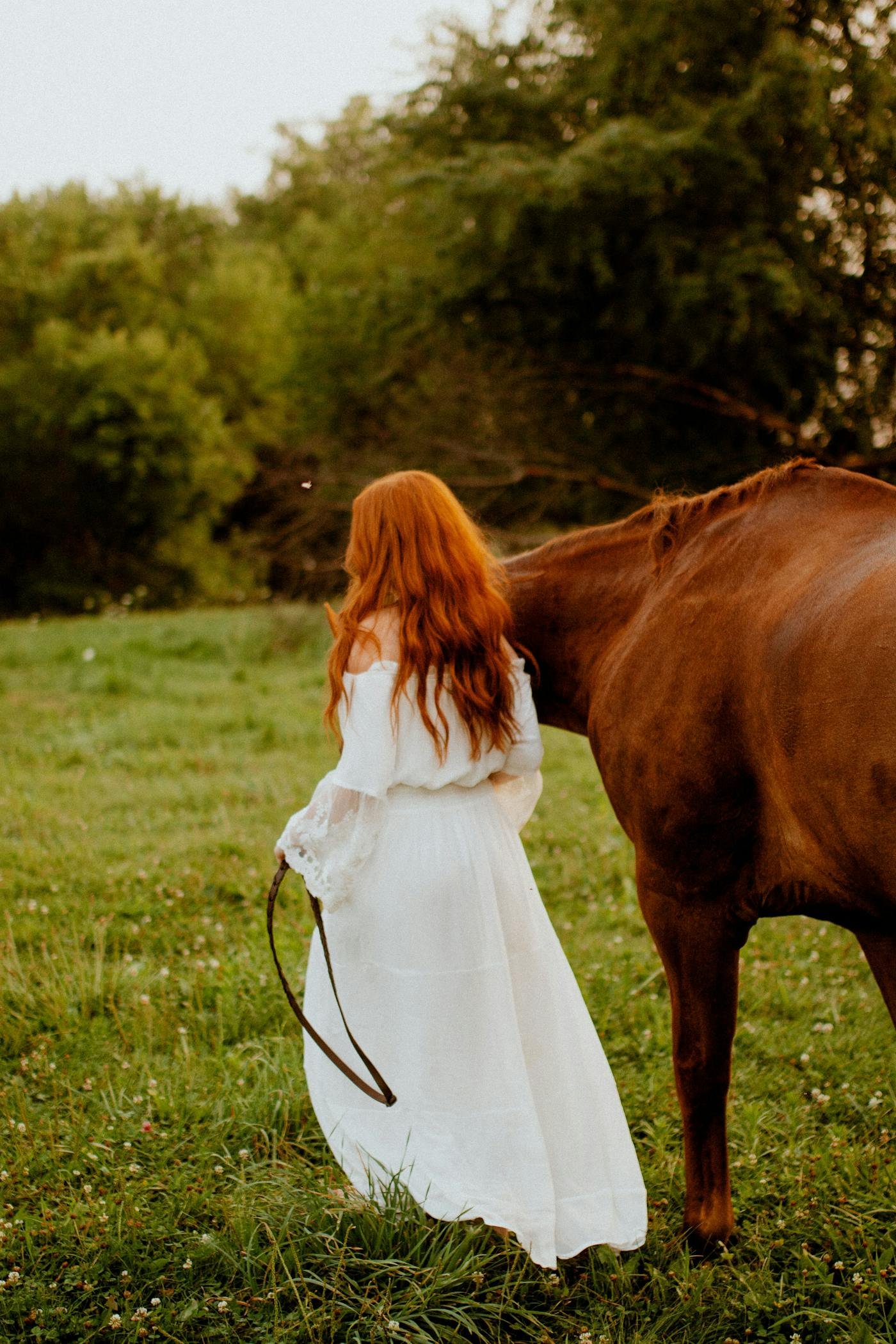 Femme en robe blanche marchant avec son cheval dans un champ verdoyant